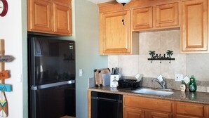 A kitchen with wooden cabinets, a black refrigerator, a sink, and a countertop with various items. - Maui Rainbows And Waves!~Gorgeous Oceanfront Views!! ~ 2 Bdrm ~Corner unit w A/C (Lahaina)