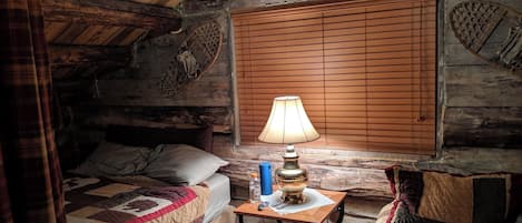 A rustic log cabin room with a bed, a table with a lamp, and a window with blinds.