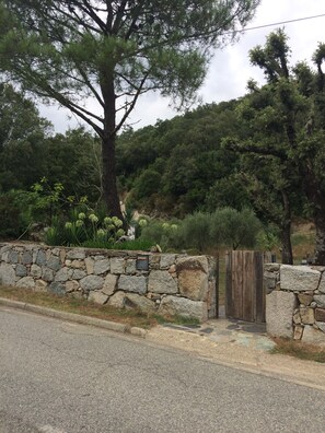 A stone wall with a wooden gate, a paved road, and a hillside with dense greenery.