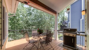 A covered outdoor patio with a round table and chairs, a grill, and a view of trees.