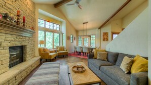 A spacious living room with a stone fireplace, comfortable seating, large windows, and a dining area.