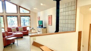 A living room with a red sofa, a small table, and a television. There are large windows and a staircase leading to another level. - Family Friendly, Air Conditioning - SHARC passes included. (Sunriver)