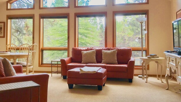 A living room with a red sofa, a dining table with chairs, and large windows offering a view of trees. - Family Friendly, Air Conditioning - SHARC passes included. (Sunriver)