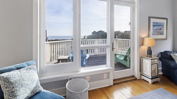 Una stanza con un'ampia finestra vista su una terrazza e il mare. Si vede un divano blu, un tavolino e una lampada su un comodino.