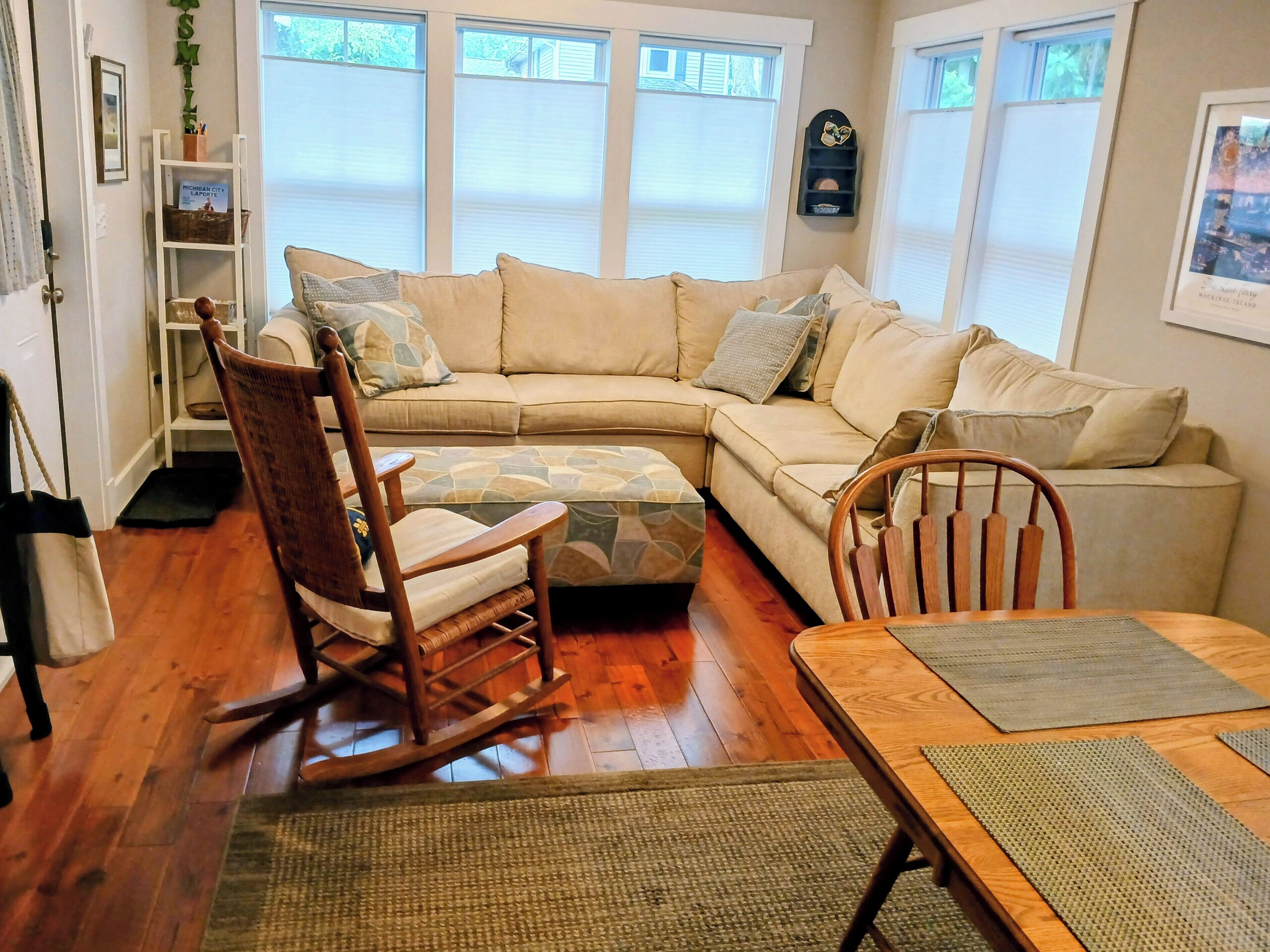 A cozy living room with a beige sectional sofa, a wooden rocking chair, and a dining table with a placemat.