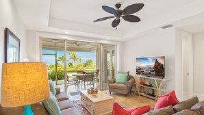 Ein geräumiges Wohnzimmer mit einer Couch, einem Couchtisch und einem Fernseher. Es gibt einen Deckenventilator, eine Lampe und einen Blick ins Freie durch eine Glastür.
