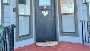 A dark grey front door with a heart-shaped decoration, flanked by two windows with shutters, and a red porch with a doormat. - Peaceful and Serene w/ Hot Tub and View (Eureka Springs)