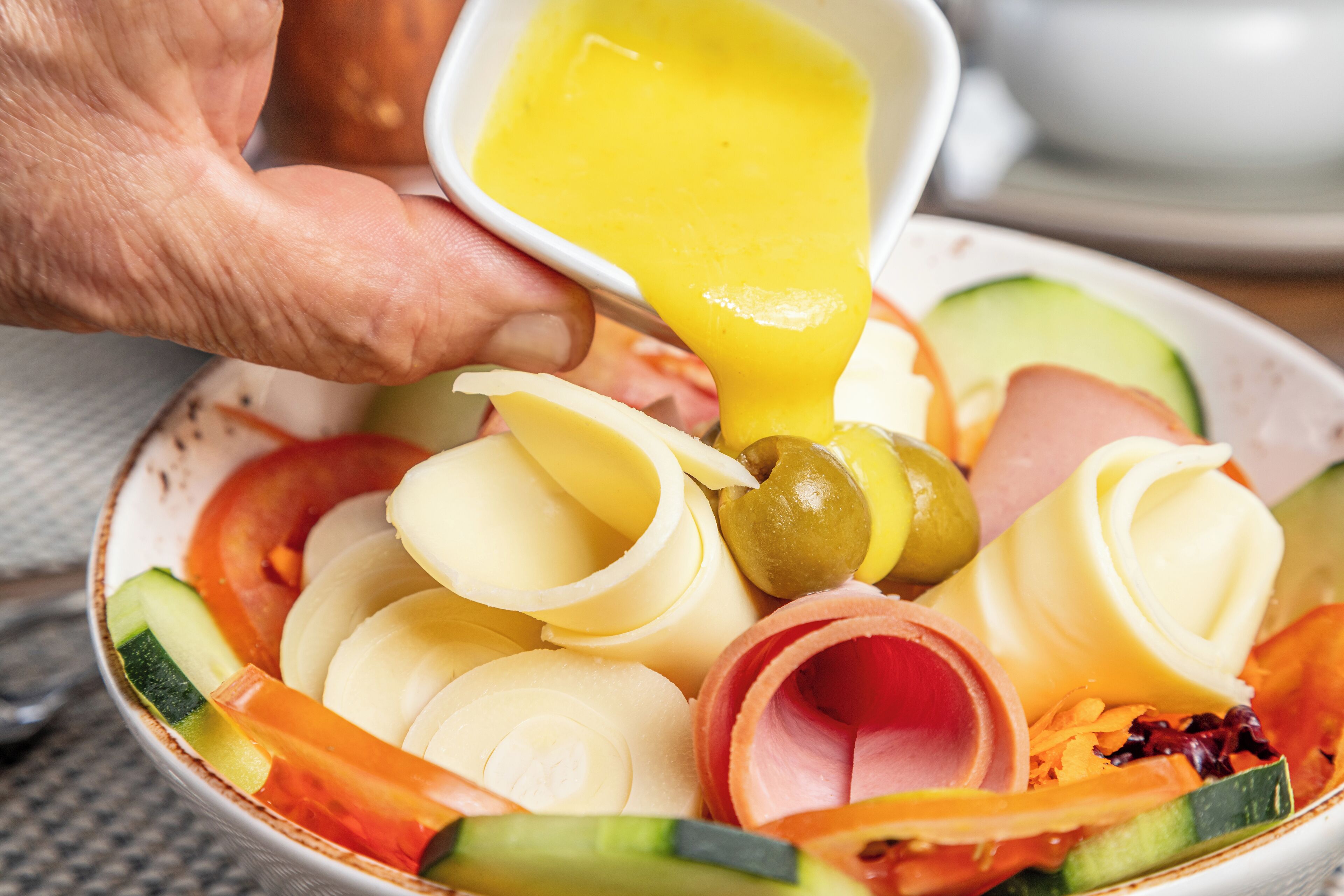 A bowl of assorted sliced vegetables and cheese with a hand pouring dressing over it.