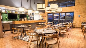 Un restaurante con suelo de madera, mesas con sillas, una zona de bar con pared verde y ventanas amplias.