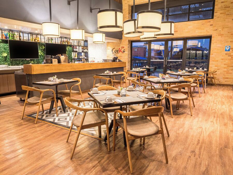 A restaurant with wooden flooring, tables with chairs, a bar area with a green wall, and large windows.