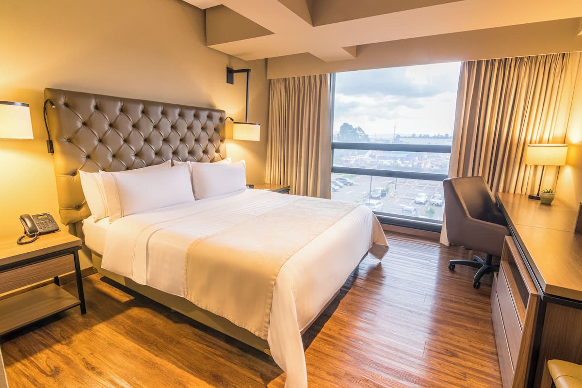 Habitación de hotel con una cama grande, un escritorio con lámpara, una silla y vistas a la ciudad a través de amplios ventanales.