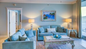 Ein Wohnzimmer mit blauem Sofagarnitur, zwei Beistelltischen mit Lampen, einem farbenfrohen Teppich und einem Bilderrahmen mit einer Küstenansicht.
