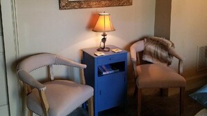 Ein Zimmer mit zwei gepolsterten Sesseln, einem blauen Beistelltisch mit Lampe, einem gerahmten Spiegel und einem Teppich mit Muster.