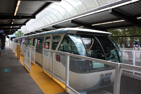 A covered train station with a modern design, featuring a yellow safety line on the platform and a metal fence separating the platform from the tracks.
