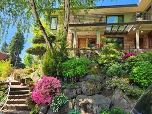 A two-story house with a stone facade, a balcony, and a well-maintained garden with various plants and flowers.