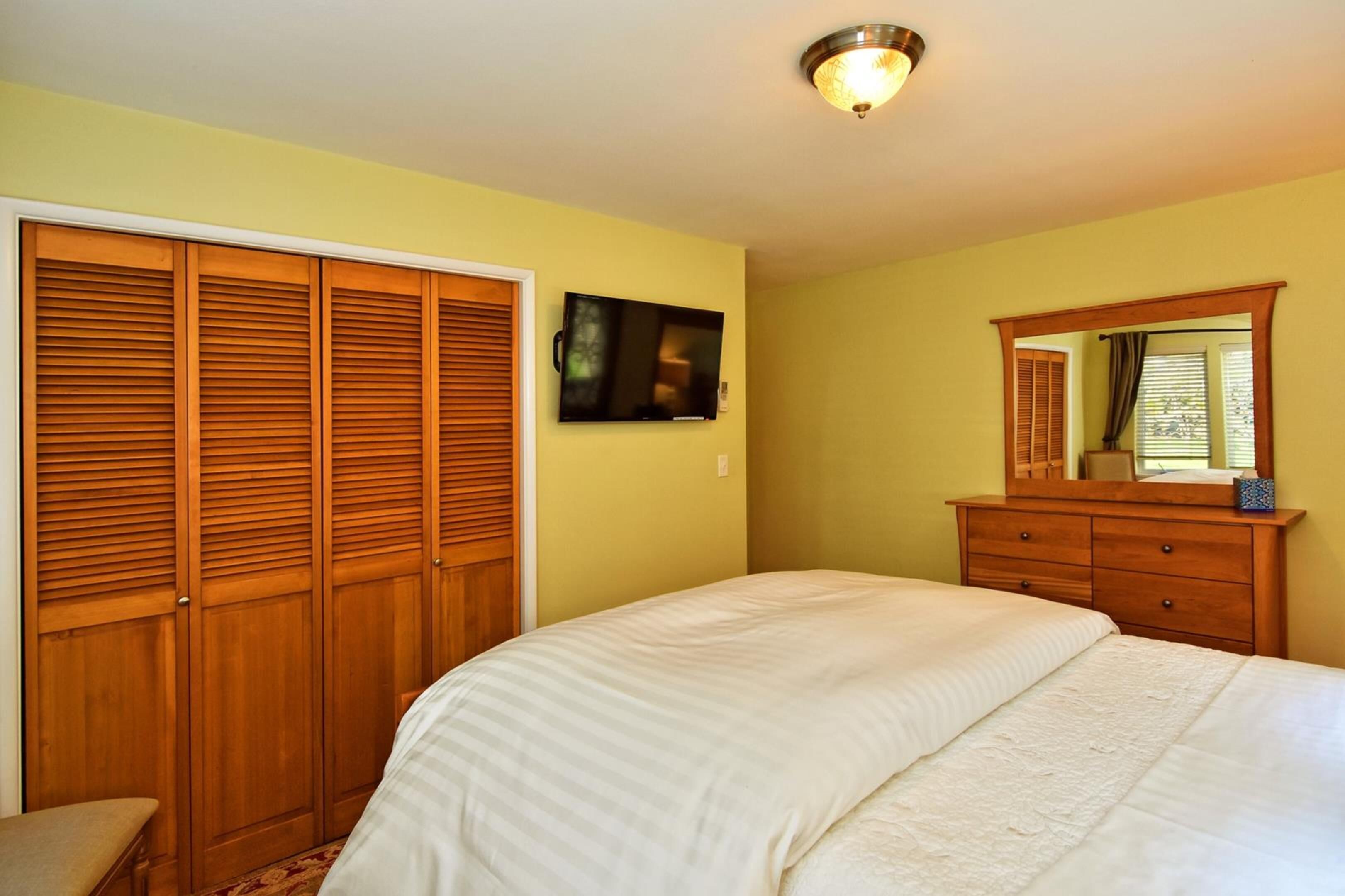 Une chambre à coucher comprenant un lit, une armoire en bois, un téléviseur à écran plat et un miroir au-dessus d’une commode.