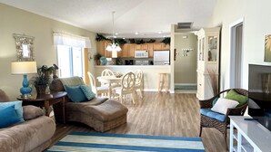 A living room with a sofa, a dining table, and chairs, a kitchen in the background, and a TV on a stand.