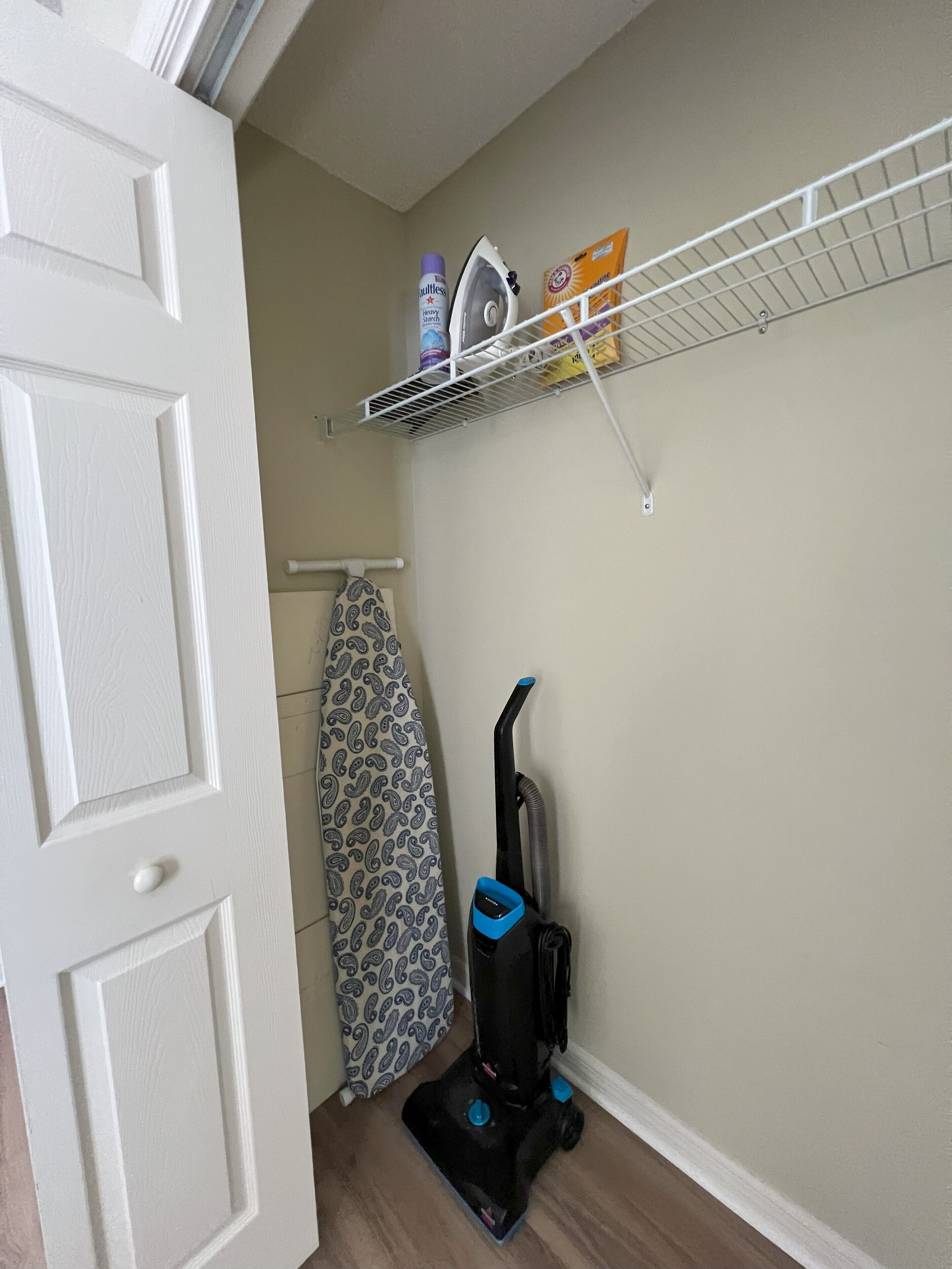 A corner of a room with a white door, a patterned ironing board, a vacuum cleaner, and a metal shelf with cleaning supplies.