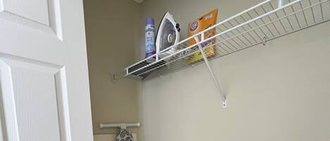 A corner of a room with a white door, a patterned ironing board, a vacuum cleaner, and a metal shelf with cleaning supplies.