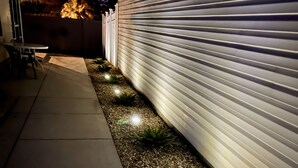 A pathway lined with small plants and pebbles, leading to a white building with a closed shutter.