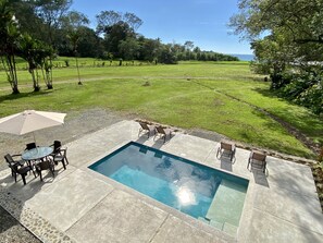 A rectangular swimming pool with a tiled deck, surrounded by lounge chairs and a table with an umbrella.