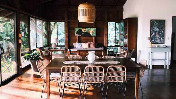 A spacious room with a wooden dining table, wicker chairs, a ceiling fan, and large windows offering a view of the outdoors. - Beach Front Home in Ballena Marine Natl Park (Uvita)