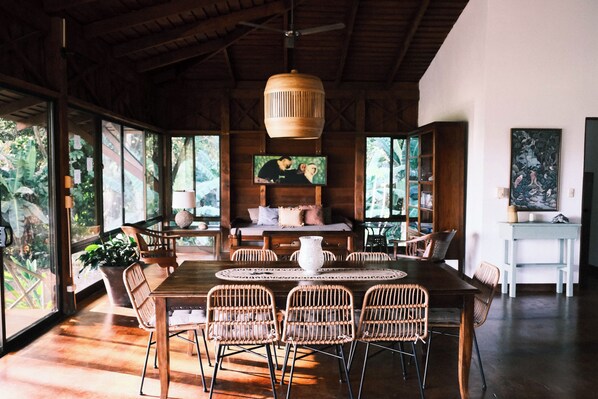 A spacious room with a wooden dining table, wicker chairs, a ceiling fan, and large windows offering a view of the outdoors.