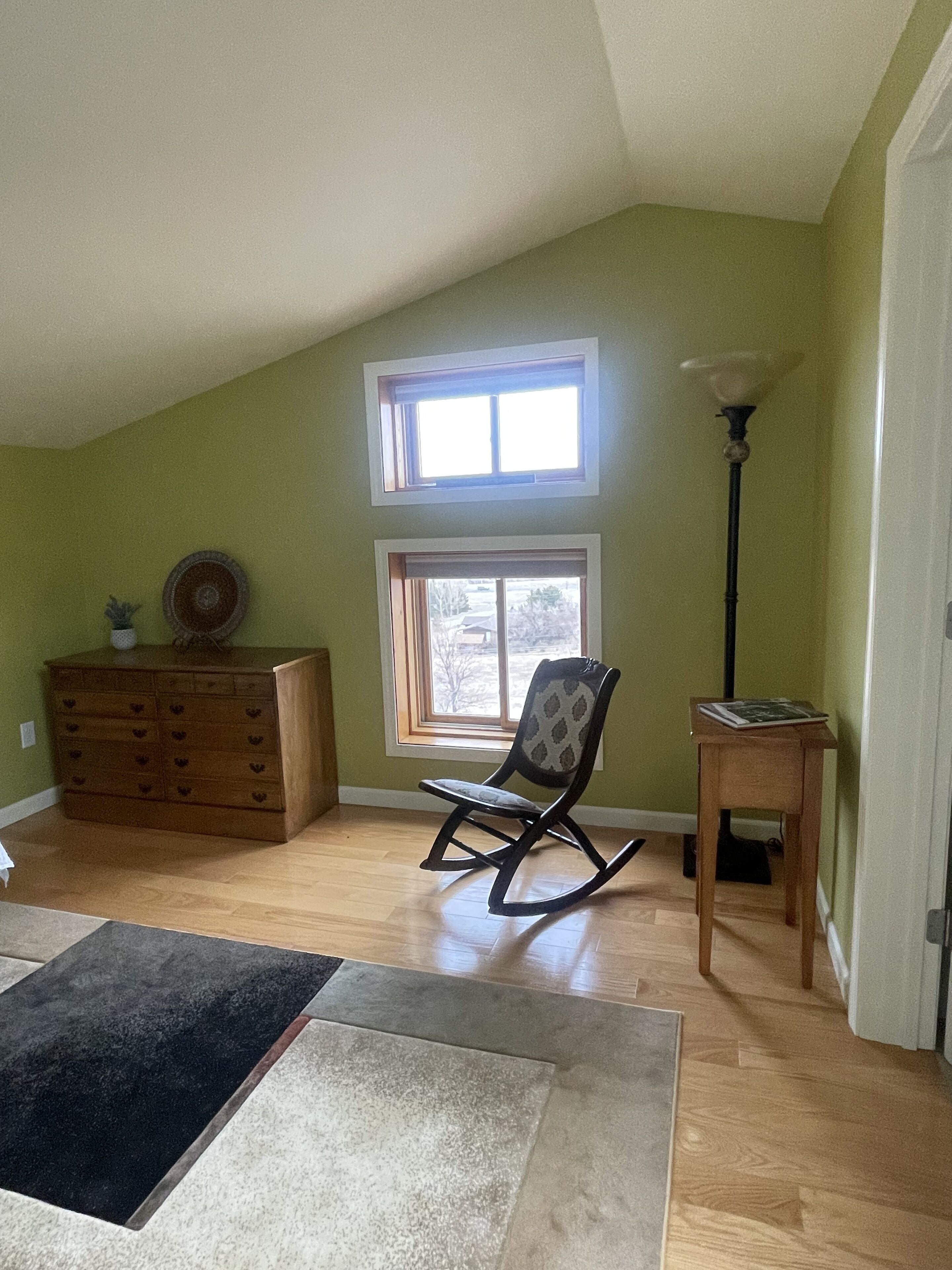 A room with a sloped ceiling, a rocking chair, a wooden dresser, a small table, and a window with a view of the outside.