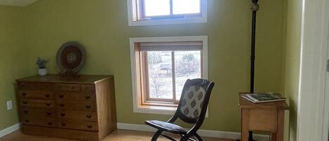 A room with a sloped ceiling, a rocking chair, a wooden dresser, a small table, and a window with a view of the outside.