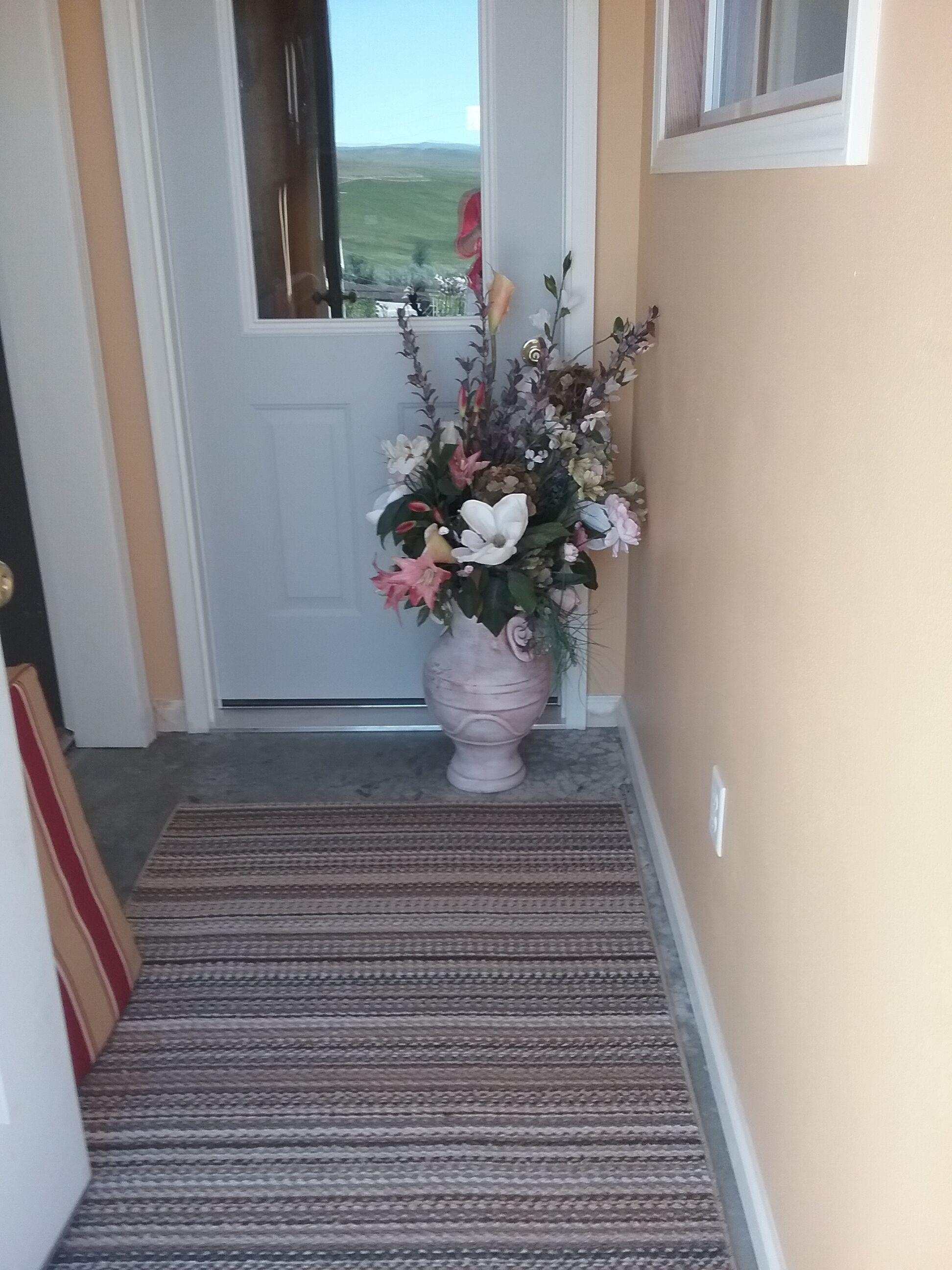 A hallway with a carpeted floor leading to a door. A large vase with flowers is placed on the floor near the door.