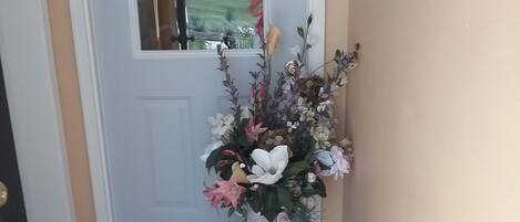 A hallway with a carpeted floor leading to a door. A large vase with flowers is placed on the floor near the door.