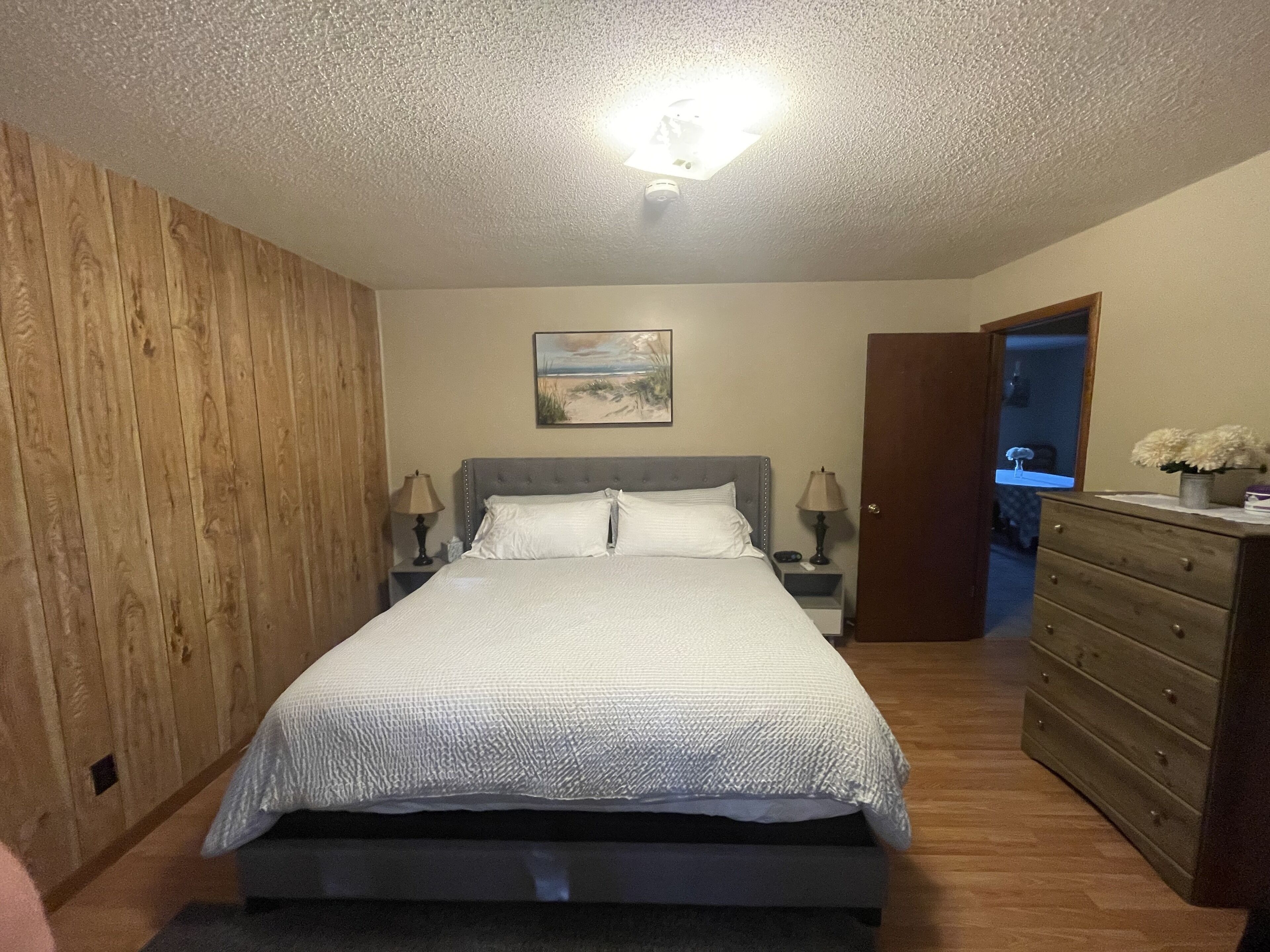 Un dormitorio con cama, mesitas de noche, cómoda, un cuadro y una puerta que da a otra habitación.
