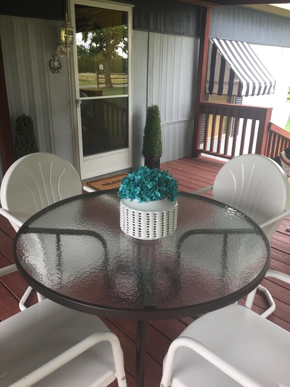 Een ronde glazen tafel met een witte mand erop, omringd door witte stoelen op een houten terras.