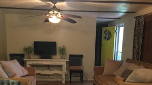 A living room with a wooden dining table, two sofas, a TV on a white stand, a ceiling fan, and a wooden floor. - Sanders Island View Beach Home only 3 blocks to the beach!  (Kingston)