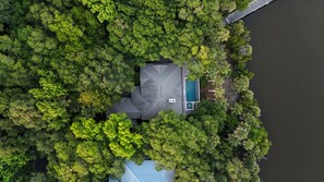 Eine Vogelperspektive zeigt ein Haus, umgeben von dichtem Baumbestand, mit einem Schwimmbecken und einem Steg.