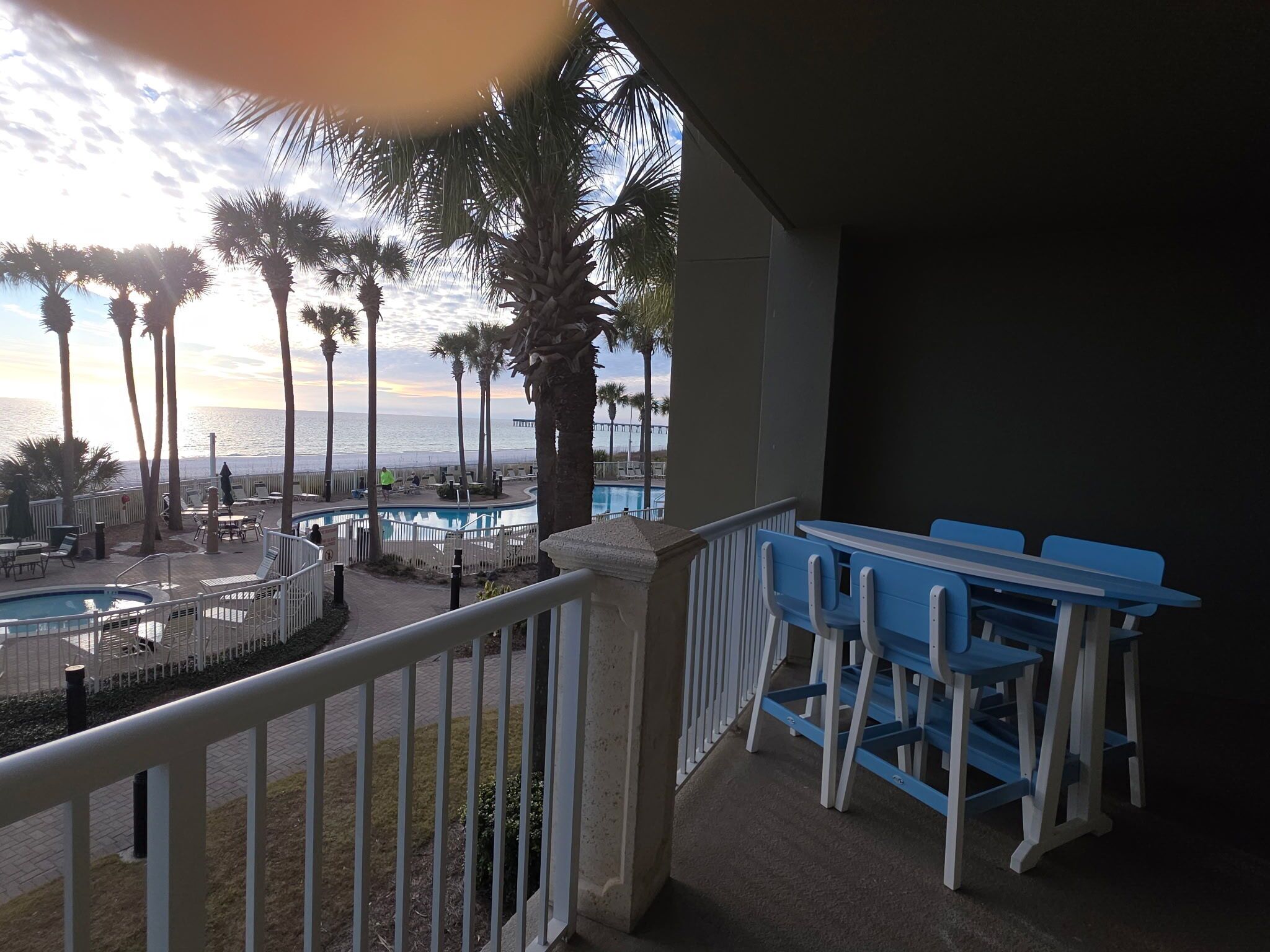 A pool area with lounge chairs and a table, surrounded by palm trees, overlooking a body of water.