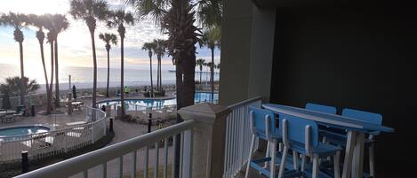 A pool area with lounge chairs and a table, surrounded by palm trees, overlooking a body of water.