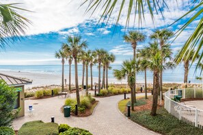 Un sendero pavimentado que conduce a una playa con palmeras, un edificio pequeño y una zona delimitada por una valla.