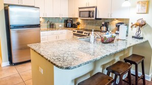 Cocina con armarios blancos, refrigerador de acero inoxidable, microondas, estufa y encimera de granito. Hay tres taburetes de bar y una lámpara colgante.