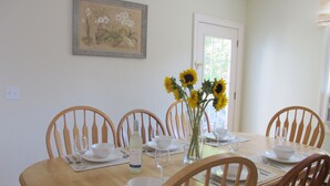 A dining room with a wooden table set for six, surrounded by six wooden chairs. There is a vase with sunflowers on the table, and a framed botanical painting on the wall. - Your search ends here! (Newport)