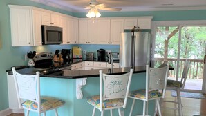 A kitchen with white cabinets, a stainless steel refrigerator, a microwave, and a coffee maker. There are three white bar stools with colorful seats. - 4 Bedroom, 2.5 Bath,  sleeps 8,  Ocean View, Large Family Room and Sunroom (Emerald Isle)