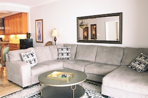 A living room with a grey sectional sofa, a coffee table, a large mirror, and a kitchen area in the background.