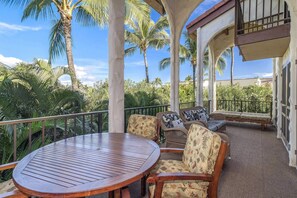 A covered outdoor seating area with a round wooden table and chairs, surrounded by palm trees and a clear sky.