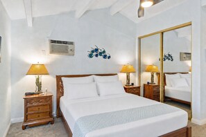 A hotel room with a large bed, two bedside tables with lamps, a wall-mounted air conditioner, and a large mirror. - Maui Hill 70 - Spacious 3BR/3BA for 8, 2 King Suites, 3 Lanais, Pool, Near Beach (Kihei)