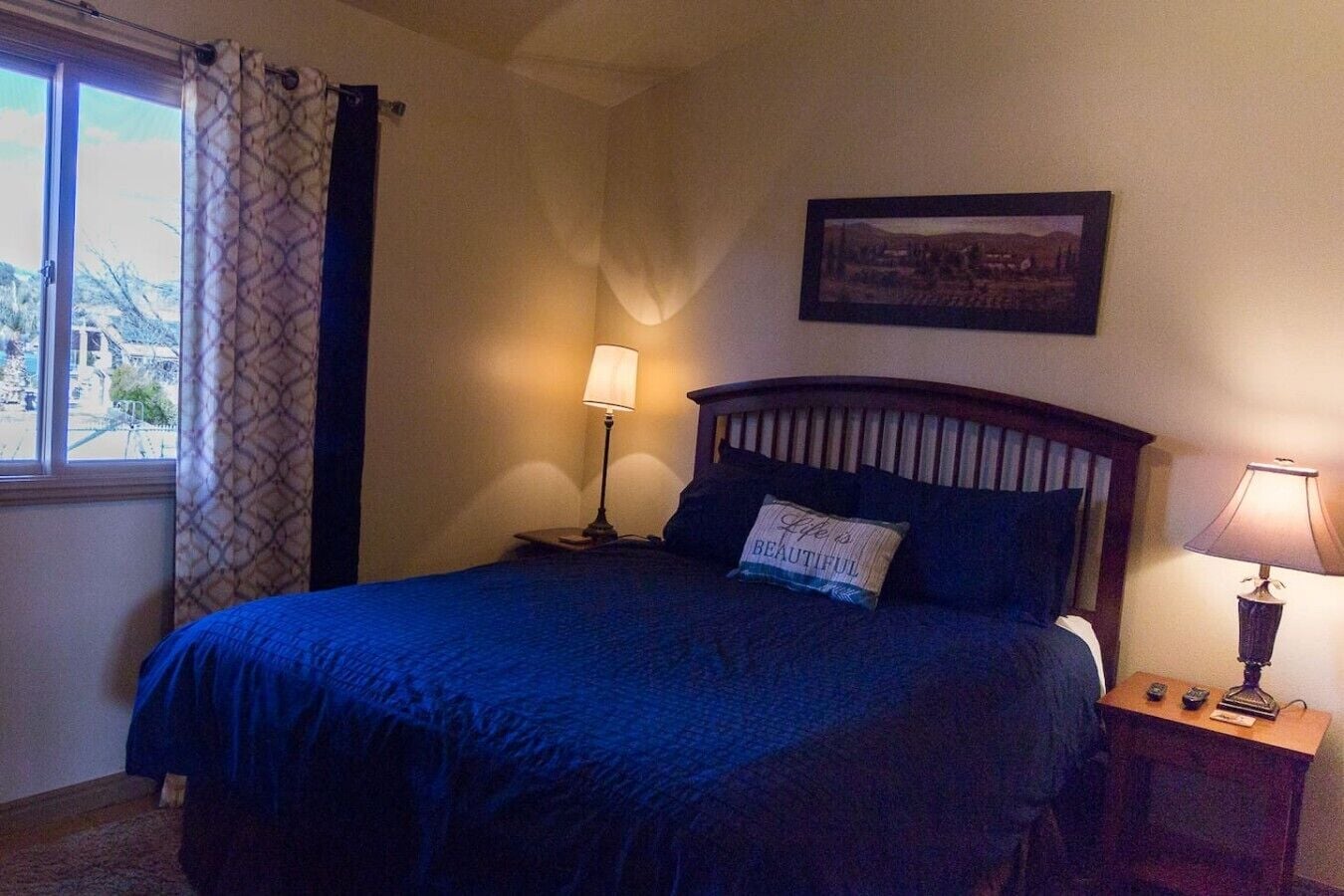 Ein Schlafzimmer mit einem Bett, zwei Nachttischlampen, einem Fenster mit Vorhängen und einem gerahmten Bild an der Wand.