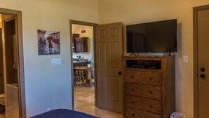 A bedroom with a bed, a dresser, a television on top, and a door leading to a kitchen area. - The Desert Hideaway, Small place, big adventures (St George)