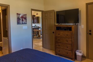 A bedroom with a bed, a dresser, a television on top, and a door leading to a kitchen area. - The Desert Hideaway, Small place, big adventures (St George)