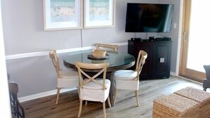 Comedor con una mesa redonda de vidrio, cuatro sillas de madera, dos cuadros con imágenes de la playa, un televisor y un mueble bajo oscuro.