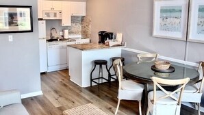 Una cocina compacta con armarios blancos, microondas y cafetera. Hay un comedor con una mesa redonda y sillas, y una pequeña isla con taburetes.