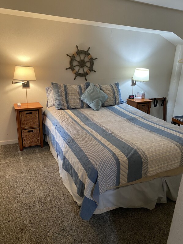 A neatly made bed with striped bedding, two bedside tables with lamps, a wall-mounted ship's wheel decoration, and a small wicker basket on one table. - Awesome oceanfront on the Sand! (La Selva Beach)